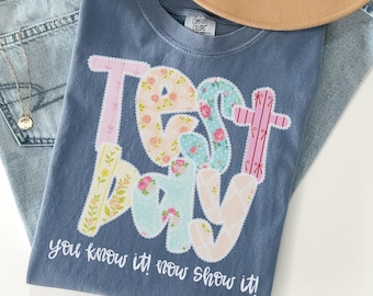 May include: A blue Comfort Colors t-shirt with the words "TEST DAY" in a floral patchwork design. The text "you know it! now show it!" is printed below the main text. A tan hat and blue jeans are in the background.