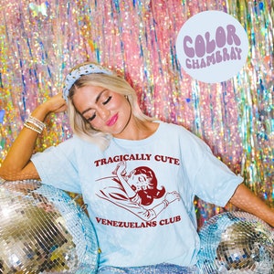 May include: A light blue t-shirt with the text "Tragically Cute" and a cartoon image of a woman lying down. The text "Venezuelans Club" is printed below the image.