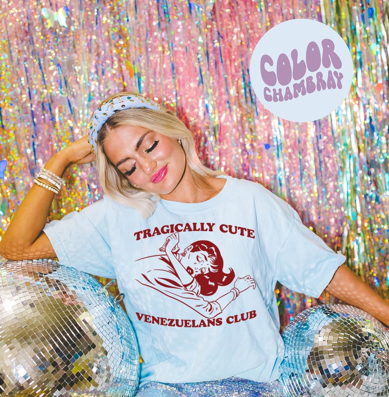 May include: A light blue t-shirt with the text "Tragically Cute" and a cartoon image of a woman lying down. The text "Venezuelans Club" is printed below the image.