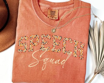 May include: A coral-colored Comfort Colors t-shirt with the words "SPEECH Squad" in a floral pattern. A gold necklace with a pendant rests on the shirt. A brown hat and a dried palm leaf are in the background.