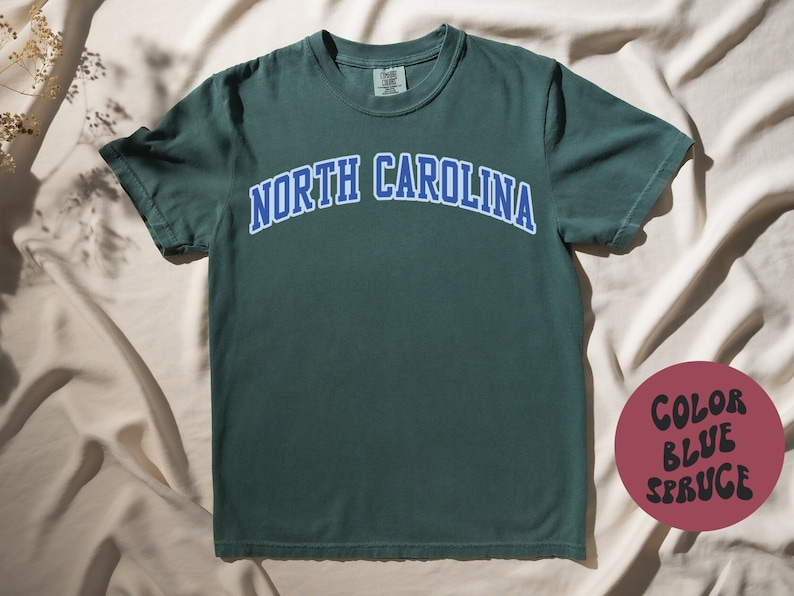 May include: A spruce-colored t-shirt with the text "NORTH CAROLINA" in blue arched lettering. The shirt is a solid color and has a crew neck. A color swatch in the lower right corner reads "COLOR BLUE SPRUCE".