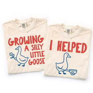 Op de afbeelding: Twee crèmekleurige t-shirts met rode tekst en blauwe illustraties. Eén shirt zegt "GROWING A SILLY LITTLE GOOSE" met een gans die een bloem vasthoudt. De andere zegt "I HELPED" met een gans die een kar trekt.