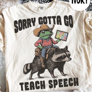 May include: Ivory t-shirt featuring a cartoon frog in a cowboy hat riding a raccoon, holding a tablet. The text on the shirt reads "SORRY GOTTA GO TEACH SPEECH". The design has a playful, illustrative style.