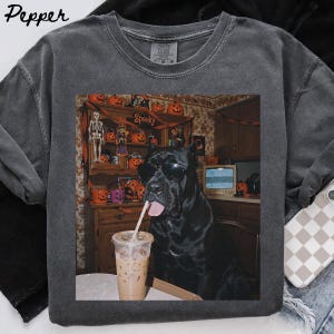 Könnte beinhalten: Ein anthrazitfarbenes T-Shirt mit einem Grafikdruck eines schwarzen Hundes mit schwarzer Sonnenbrille, der neben einem Eiskaffee mit Strohhalm sitzt. Der Hintergrund zeigt eine Küchenanrichte, die mit Kürbissen und einem Skelett dekoriert ist.