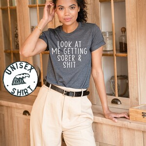 May include: A dark gray t-shirt with the text "LOOK AT ME GETTING SOBER & SHIT" printed in white.