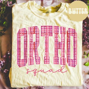 May include: A pale yellow t-shirt featuring the word "ORTHO" in large, pink plaid letters and the word "squad" in cursive below. The word "BUTTER" is in a circle in the upper right corner.