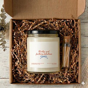 Könnte beinhalten: Geschenkbox mit einem Glas-Kerzenglas mit schwarzem Deckel, einem kleinen Glasröhrchen mit Streichhölzern und braunem Papierfüllmaterial. Das Kerzenetikett lautet "thirty and fucking fabulous" in Rot und Blau.