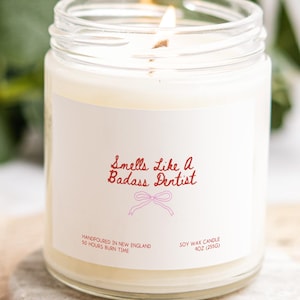 A white candle in a glass jar with a label that reads "Smells like a Badass Dentist".
