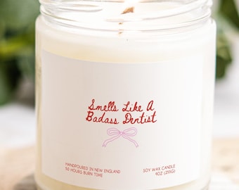May include: A lit soy wax candle in a clear glass jar. The label reads "Smells Like a Badass Dentist" in red script. The candle is hand-poured in New England and has a 50-hour burn time. The candle weighs 9oz (255g).