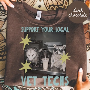 以下が含まれることがあります： ダークチョコレートブラウンのクルーネックトレーナー。ライトブルーで「SUPPORT YOUR LOCAL VET TECHS」の文字がプリントされています。グラフィックには、サングラスと聴診器を身につけたオポッサム、スカンク、アライグマのモノクロ写真が描かれています。