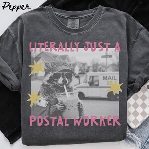Könnte beinhalten: Ein anthrazitfarbenes Kurzarm-T-Shirt mit einer schwarz-weißen Grafik eines Waschbären, der eine Mütze und eine Sonnenbrille trägt und ein Getränk hält. Der Text LITERALLY JUST A POSTAL WORKER ist in Pink über und unter dem Bild aufgedruckt.