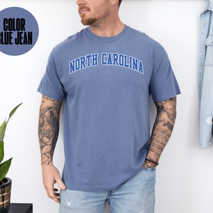 May include: Blue jean colored t-shirt with the text "NORTH CAROLINA" in a blue arched design. The shirt is short-sleeved and appears to be made of a soft, comfortable material. The color is indicated in a circle in the upper left corner.