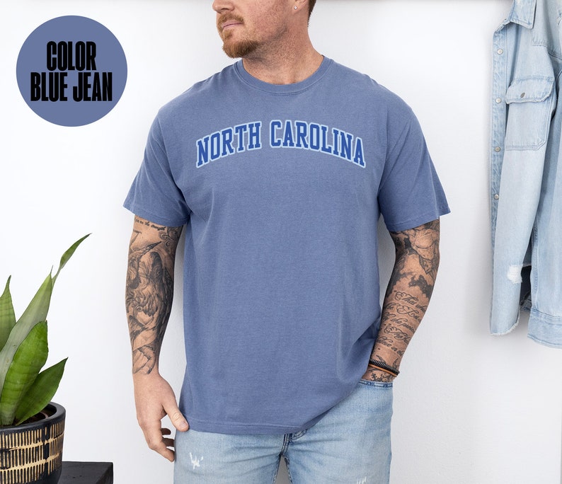 May include: Blue jean colored t-shirt with the text "NORTH CAROLINA" in a blue arched design. The shirt is short-sleeved and appears to be made of a soft, comfortable material. The color is indicated in a circle in the upper left corner.