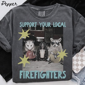 以下が含まれることがあります： 消防車の前に3匹の動物が描かれたチャコールグレーのTシャツ。テキストは「SUPPORT YOUR LOCAL FIREFIGHTERS」と書かれています。動物は赤いヘルメットをかぶったオポッサム、飲み物を持ったスカンク、サングラスをかけたアライグマです。