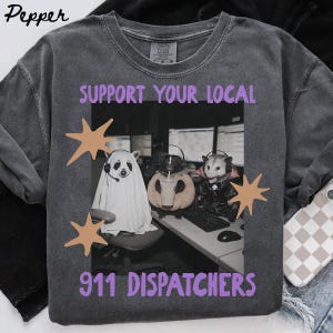 Può includere: Una t-shirt grigio antracite con una grafica di tre opossum che indossano cuffie a una scrivania. Il testo recita SUPPORT YOUR LOCAL 911 DISPATCHERS in viola. Gli opossum sono vestiti da fantasma, zucca e vampiro, con accenti a forma di stella.