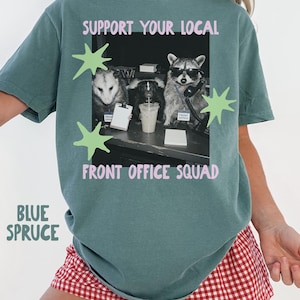 May include: A blue spruce t-shirt featuring a graphic of a possum, a skunk, and a raccoon in an office setting. The text reads SUPPORT YOUR LOCAL FRONT OFFICE SQUAD in pink letters. Light green star shapes surround the central black and white image.