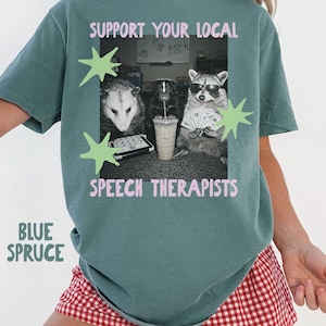 Könnte beinhalten: Ein blau-fichtenfarbenes T-Shirt mit einer Grafik aus Opossum, Stinktier und Waschbär sowie dem rosa Schriftzug SUPPORT YOUR LOCAL SPEECH THERAPISTS. Die Tiere sind mit einer Kommunikationstafel, einem Getränk und einer Sonnenbrille dargestellt.