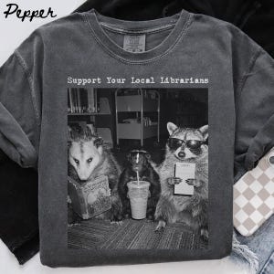Puede incluir: Una camiseta de cuello redondo gris carbón con un gráfico en blanco y negro de una zarigüeya leyendo un libro, una mofeta sosteniendo una bebida y un mapache con gafas de sol sosteniendo un carné de biblioteca. El texto dice Support Your Local Librarians.