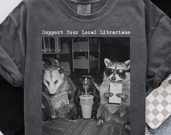 Peut inclure: Un t-shirt à col rond gris anthracite avec un graphique en noir et blanc représentant un opossum lisant un livre, une mouffette tenant une boisson et un raton laveur portant des lunettes de soleil et une carte de bibliothèque. Texte : Support Your Local Librarians.