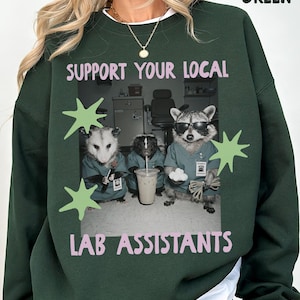Może przedstawiać: Leśno-zielona bluza z okrągłym dekoltem z nadrukiem trzech zwierząt ubranych w turkusowe fartuchy laboratoryjne. Napis brzmi "SUPPORT YOUR LOCAL LAB ASSISTANTS" w jasnoróżowym kolorze. Zwierzęta to opos, skunks i szop pracz w okularach przeciwsłonecznych.