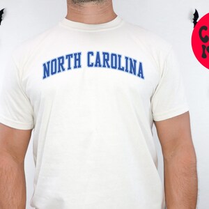 May include: Ivory-colored t-shirt with the text "NORTH CAROLINA" in blue, arched across the chest. The shirt is short-sleeved. A red circle with the words "COLOR IVORY" is visible. Black bat decorations are on the white background.