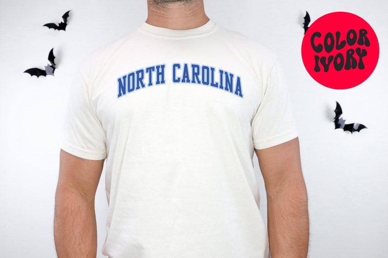 May include: Ivory-colored t-shirt with the text "NORTH CAROLINA" in blue, arched across the chest. The shirt is short-sleeved. A red circle with the words "COLOR IVORY" is visible. Black bat decorations are on the white background.