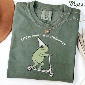 May include: A moss green t-shirt featuring the text "Off to commit mathfoolery" above a cartoon frog on a scooter, wearing a wizard hat. A gold necklace and a brown hat are also visible.