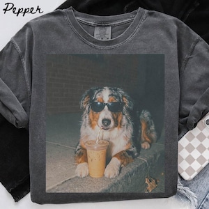 Puede incluir: Una camiseta de cuello redondo gris carbón con un estampado gráfico de un perro pastor australiano que lleva gafas de sol negras y sostiene un vaso de plástico transparente con un café helado y una pajita.