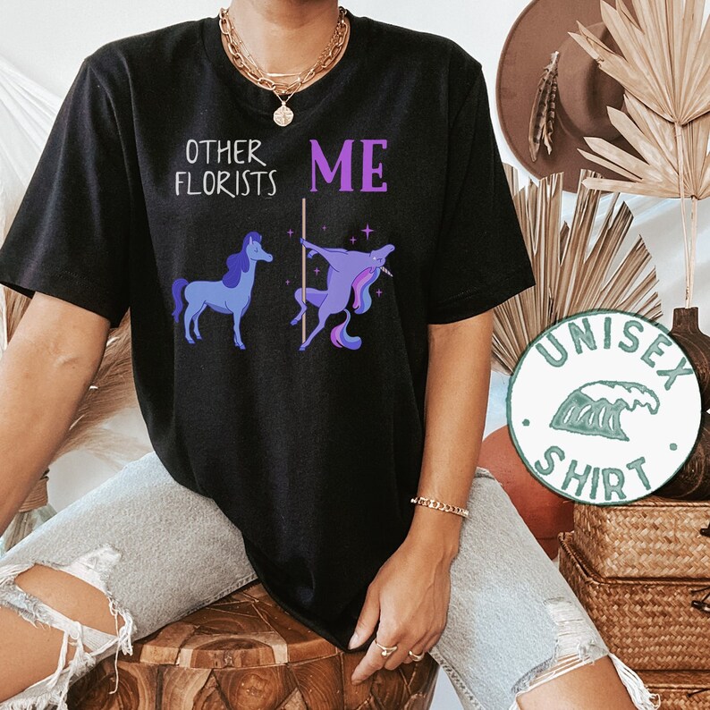 May include: Black t-shirt with a graphic of two unicorns. One unicorn is standing normally, while the other is on a pole. The text "Other Florists" is above the normal unicorn, and the text "ME" is above the unicorn on the pole. The shirt is labeled "Unisex Shirt".