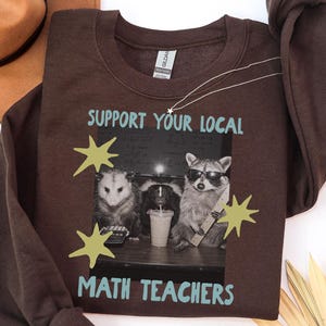 Op de afbeelding: Een donkerchocoladebruin sweatshirt met ronde hals en een grafische print van een opossum, een stinkdier en een wasbeer aan een bureau. De tekst SUPPORT YOUR LOCAL MATH TEACHERS is lichtblauw, geaccentueerd door vier gele sterren.