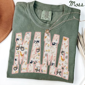 a green t-shirt with a large word "MAMA" printed on it, surrounded by various farm-themed illustrations such as tractors, animals, and flowers.