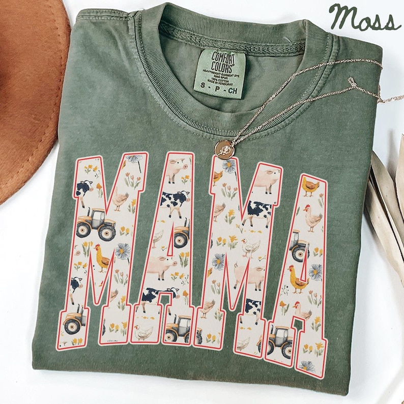 a green t-shirt with a large word "MAMA" printed on it, surrounded by various farm-themed illustrations such as tractors, animals, and flowers.