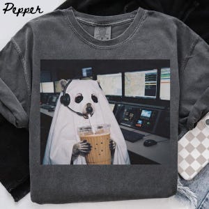Pode incluir: Uma t-shirt de gola redonda cinzento-carvão com uma estampa gráfica de um guaxinim vestido com um lençol de fantasma branco, auscultadores pretos e a segurar um copo de plástico com palhinha. O fundo mostra um escritório escuro com monitores.