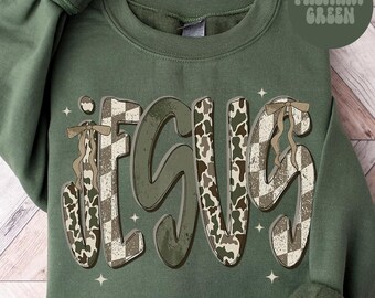 May include: Military green sweatshirt with the word "JESUS" in a distressed, layered design. The letters feature a mix of camouflage and checkered patterns, accented with decorative bows. The sweatshirt is a solid military green color.
