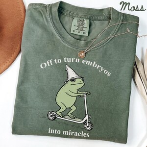 以下が含まれることがあります： モスグリーンのTシャツ。テキストは「Off to turn embryos into miracles」です。ウィザードハットをかぶったカエルの漫画がスクーターに乗っています。金のネックレスと茶色の帽子も表示されています。