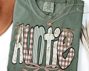 May include: A moss green Comfort Colors t-shirt with the word "Auntie" in a decorative font. The letters have a plaid, striped, and duck pattern. A gold necklace and a brown bow are also visible.