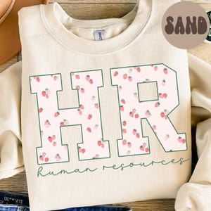 Peut inclure: Sweat-shirt couleur sable avec les lettres "HR" bordées de vert, remplies d'un motif de fraises. Les mots "human resources" sont écrits en vert en dessous. Un cercle "SAND" est dans le coin supérieur droit.