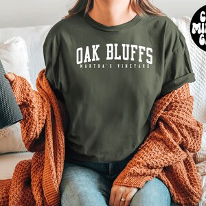 May include: A green t-shirt with the text "Oak Bluffs Martha's Vineyard" in white lettering. The person is wearing a brown cardigan and blue jeans.
