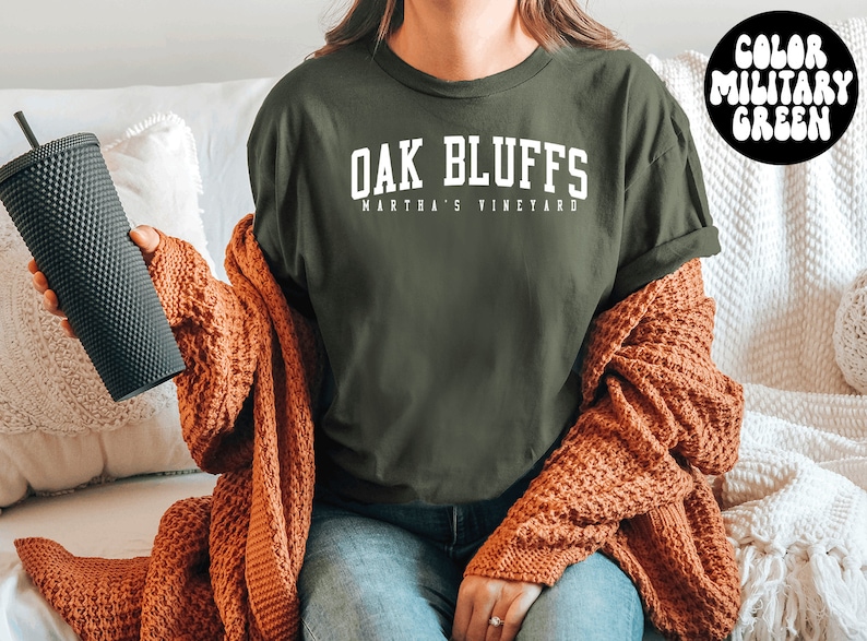 May include: A green t-shirt with the text "Oak Bluffs Martha's Vineyard" in white lettering. The person is wearing a brown cardigan and blue jeans.