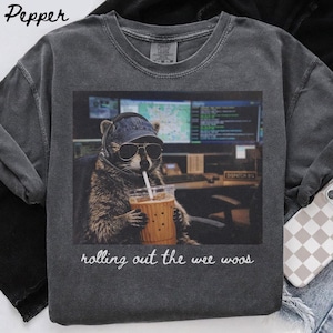 Puede incluir: Sudadera gris oscuro de cuello redondo con un gráfico de un mapache con gafas de sol, gorra vaquera y auriculares, bebiendo una bebida. El texto "rolling out the wee woos" está impreso debajo del gráfico.