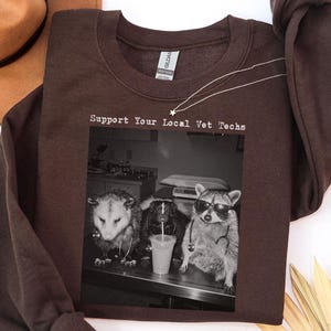 Könnte beinhalten: Ein dunkel schokoladenbraunes Langarm-Sweatshirt mit einem schwarz-weißen Grafikdruck eines Opossums, eines Stinktiers und eines Waschbären, die Stethoskope und Sonnenbrillen tragen, mit dem Text Support Your Local Vet Techs über dem Bild.