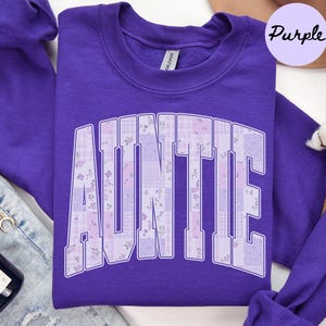 May include: A purple sweatshirt with the word "AUNTIE" in large, patchwork-style letters. The letters feature floral and plaid patterns in shades of purple and white. The sweatshirt has a crew neck and long sleeves.