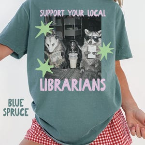 Puede incluir: Una camiseta color abeto azul con un gráfico de un oposum, una mofeta y un mapache en una biblioteca. El texto SUPPORT YOUR LOCAL LIBRARIANS aparece en letras rosas. El diseño incluye estrellas de color verde claro y una foto en blanco y negro.