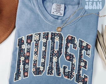 May include: A blue jean Comfort Colors t-shirt with the word "NURSE" in a floral pattern. The shirt has a crew neck and short sleeves. A gold necklace with a pendant is draped on the shirt.