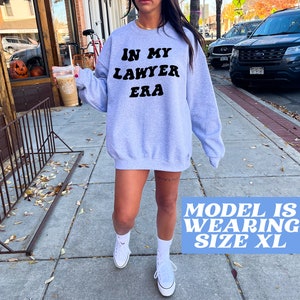 May include: A gray sweatshirt with the text "In My Lawyer Era" in black retro-style lettering. The model is wearing size XL.