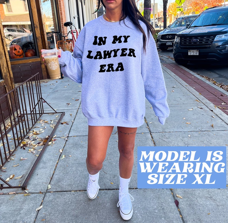 May include: A gray sweatshirt with the text "In My Lawyer Era" in black retro-style lettering. The model is wearing size XL.