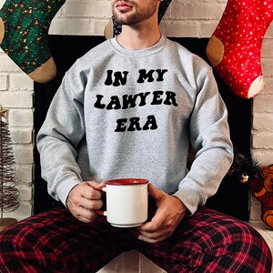 May include: A gray sweatshirt with the text "IN MY LAWYER ERA" in black retro-style lettering. The person wearing the sweatshirt is holding a white mug with a red rim.