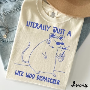 Puede incluir: Una camiseta color marfil con un gráfico de líneas azules de un ratón con gafas de sol y auriculares mientras sostiene una bebida. El texto LITERALLY JUST A WEE WOO DISPATCHER aparece en letras azules. Etiqueta de cuello Comfort Colors.