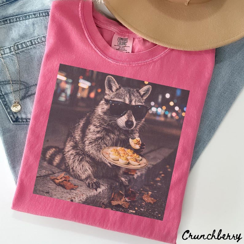 May include: A bright pink t-shirt featuring a graphic print of a raccoon wearing black sunglasses and holding a plate of deviled eggs. The shirt has a crew neckline and a Comfort Colors label on the inside collar. The fabric is a solid pink color.