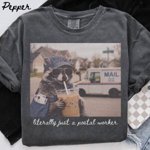 May include: A charcoal grey t-shirt featuring a graphic of a raccoon wearing a blue baseball cap and sunglasses, holding a plastic cup with a straw. The raccoon carries a blue mail bag. Below the image, the text reads, literally just a postal worker.